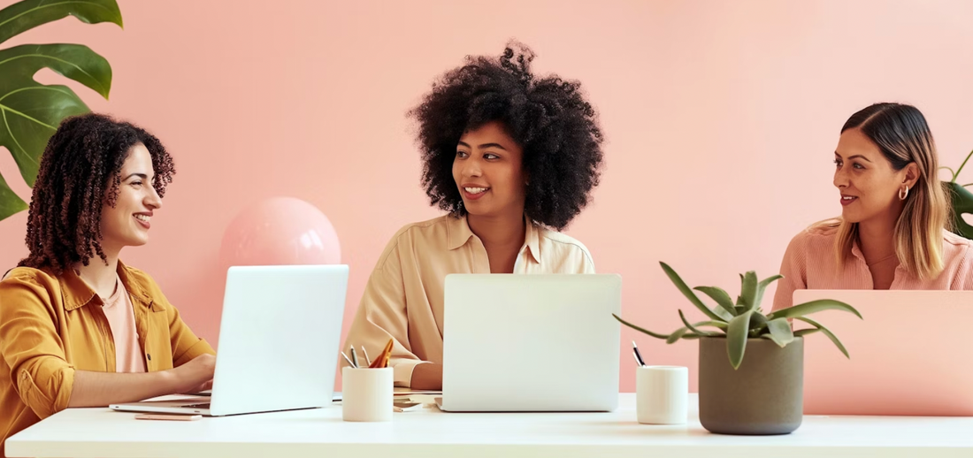 Three diverse female entrepreneurs engaging in a collaborative discussion at a modern workspace, with laptops and a plant on the table