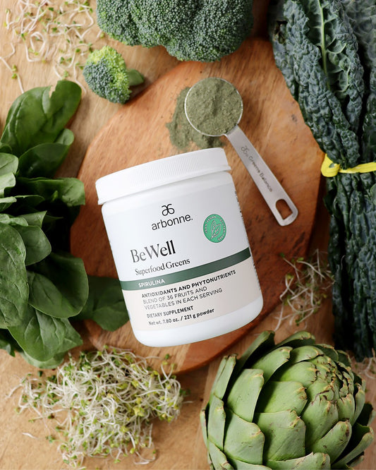 Arbonne BeWell Superfood Greens container surrounded by fresh vegetables, spinach, broccoli, kale, and a measured scoop of green powder.