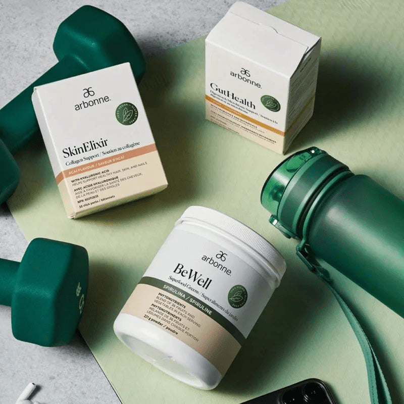 Arbonne SkinElixir, GutHealth, and BeWell Superfood Greens products displayed with green dumbbells and a water bottle.