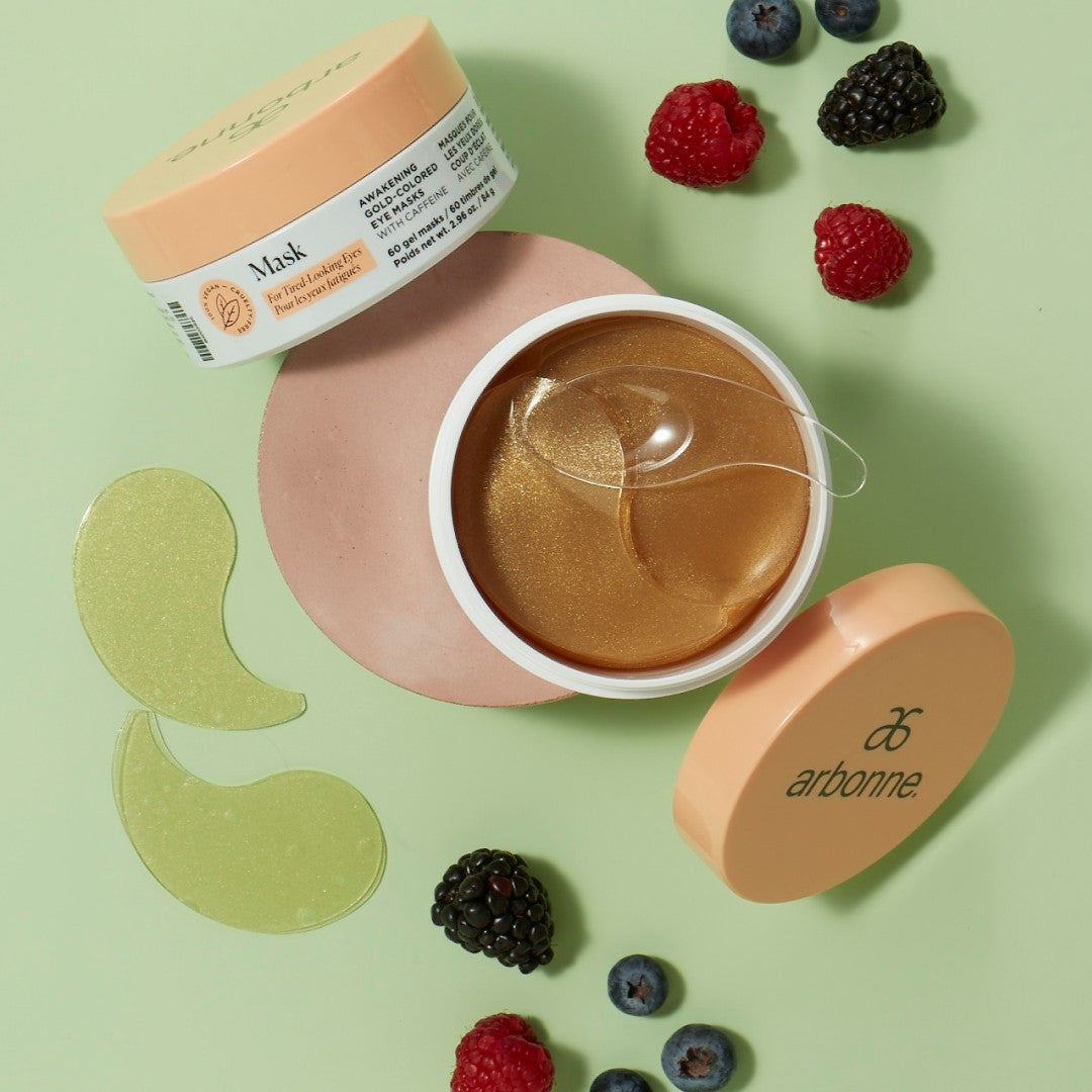 Arbonne eye mask patches displayed with fresh berries on a light green background. The image shows an open jar of golden eye mask patches, with the lid and packaging visible.