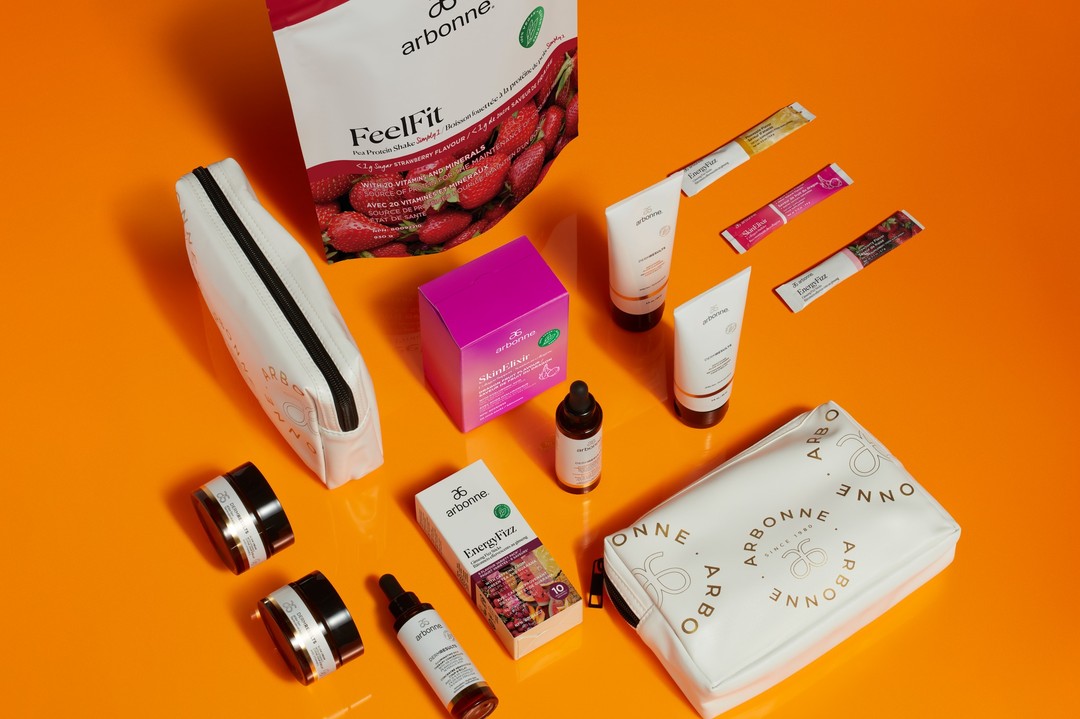 Collection of Arbonne products including FeelFit protein shake, EnergyFizz sticks, SkinElixir, skincare creams, serums, and white cosmetic bags displayed on an orange background.