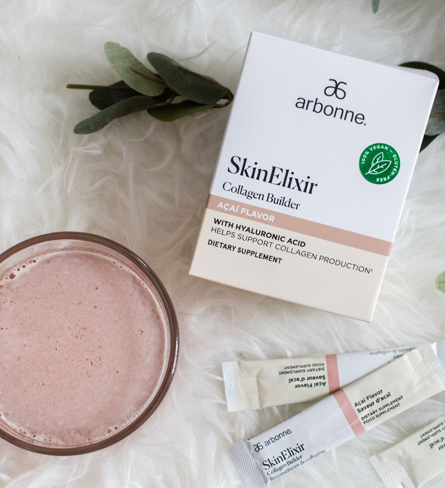 Arbonne SkinElixir Collagen Builder in Acai Flavor with a prepared drink and sachets on a plush white background.