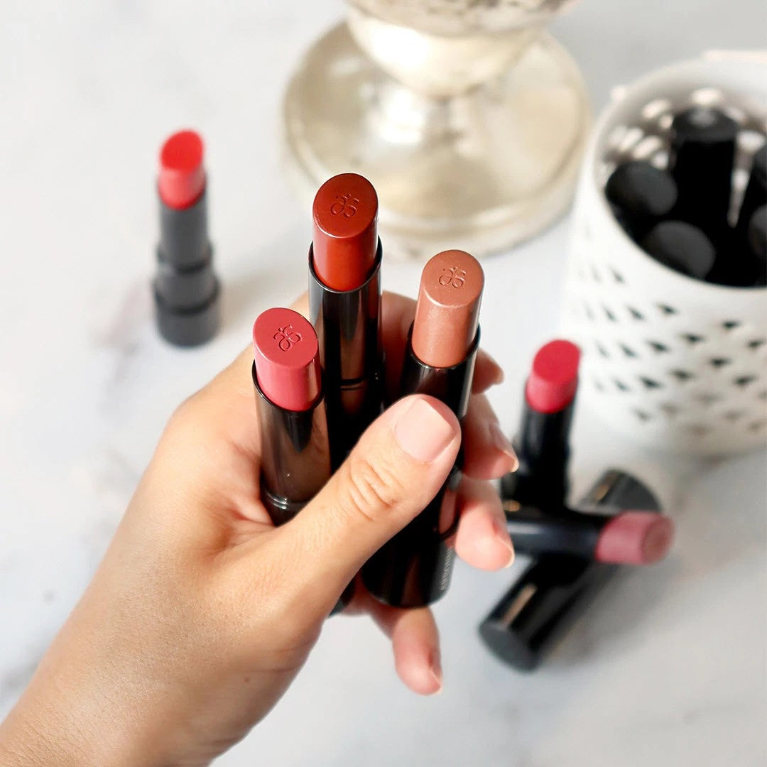 Hand holding four Arbonne lipsticks in various shades, with additional lipsticks scattered on a marble surface and in a white holder in the background.