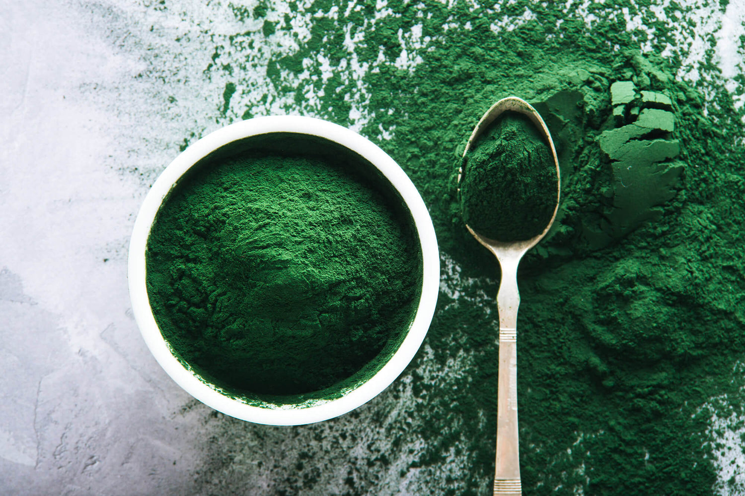 Bowl of vibrant green spirulina powder next to a spoonful of spirulina powder on a textured surface.