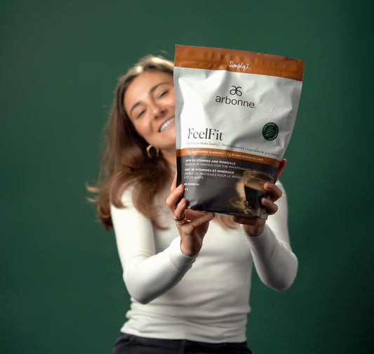 Woman holding a bag of Arbonne FeelFit protein shake powder in coffee flavour against a green background.