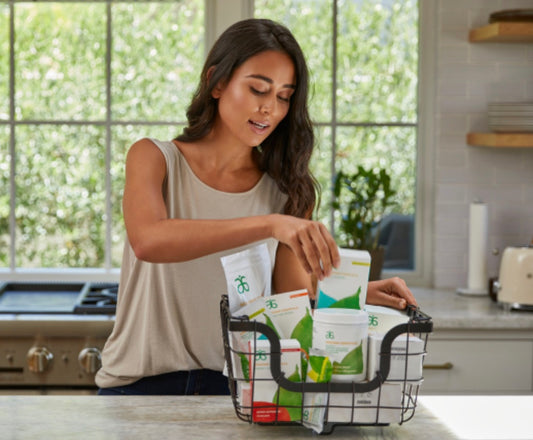 Woman organizing Arbonne wellness products in a kitchen, selecting items from a basket filled with various health supplements in front of a window overlooking a green yard.
