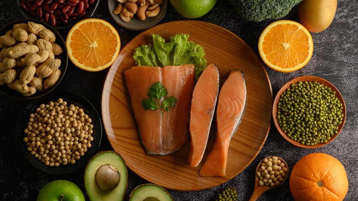 Assortment of plant-based foods including avocado, peanuts, yellow peas, green peas, oranges, and salmon arranged on a wooden plate..
