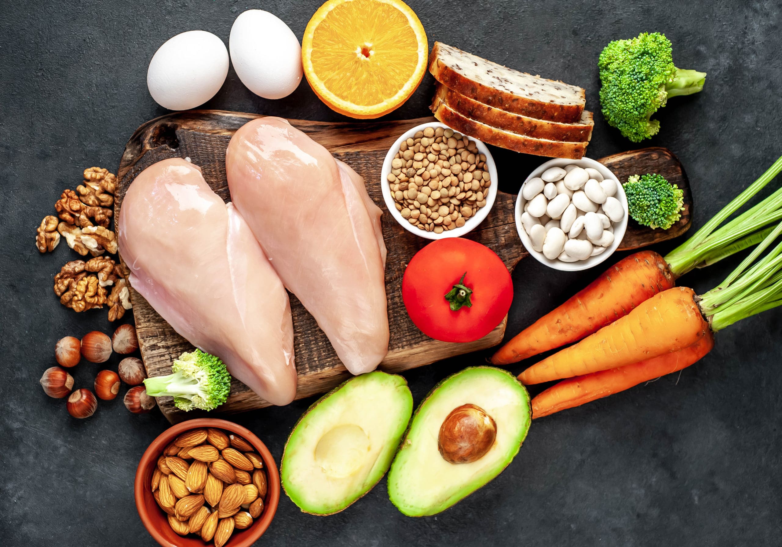 Variety of healthy foods including raw chicken breast, eggs, orange slice, bread, lentils, white beans, tomato, carrots, broccoli, avocado, almonds, walnuts, and hazelnuts on a dark surface.