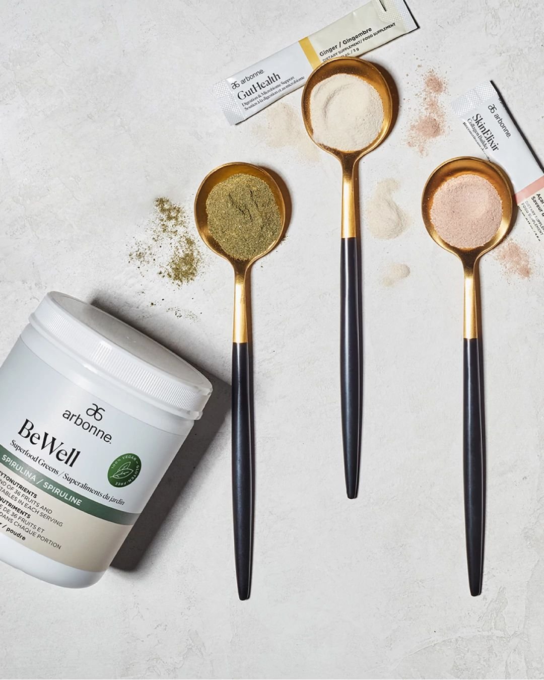 Arbonne BeWell superfood greens with GutHealth and SkinElixir supplements in measuring spoons on a textured surface.