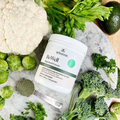 Arbonne BeWell Superfood Greens powder surrounded by fresh vegetables including cauliflower, Brussels sprouts, celery, and broccoli.