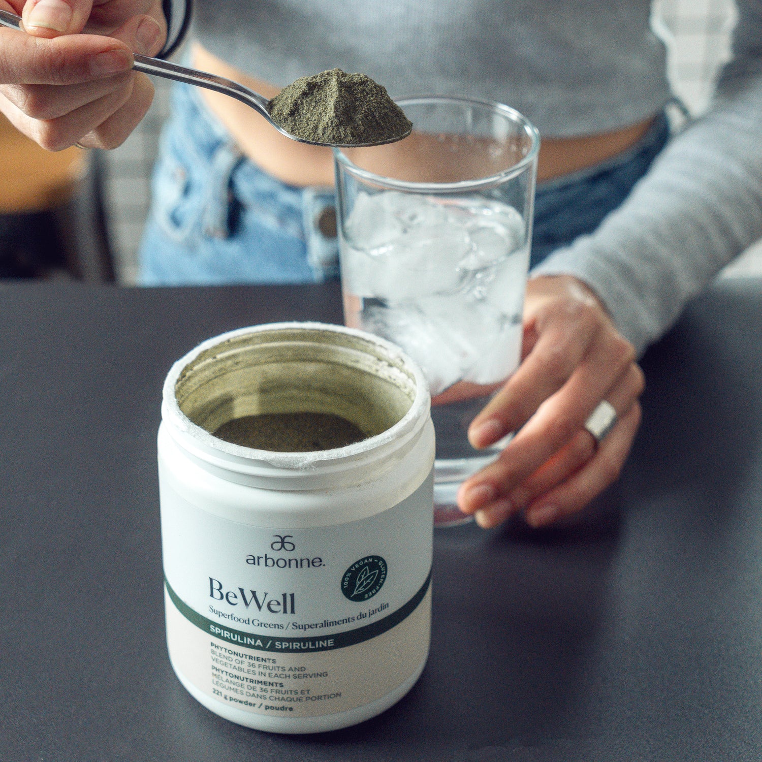 Person scooping green BeWell superfood powder from an Arbonne container, with a glass of ice water in the background.