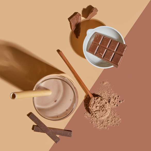 Elevated view of Arbonne chocolate protein shake with bamboo straw, powder, and dark chocolate pieces on a dual-tone background.