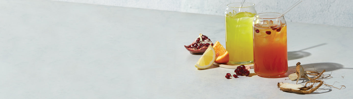 Two glasses of Arbonne Energy Fizz Sticks beverages, one yellow and one orange with a gradient effect, surrounded by fresh citrus fruits and pomegranate on a modern kitchen counter.