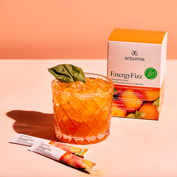 Arbonne EnergyFizz Ginseng Fizz Sticks next to a glass of the prepared energy drink with a leaf garnish on a coral background.
