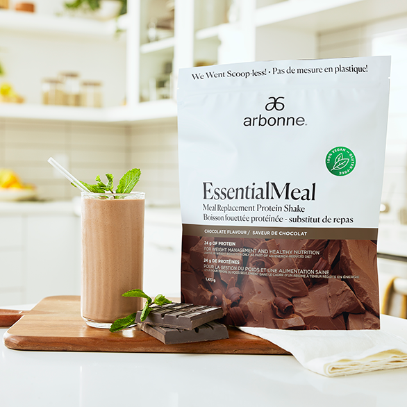 Chocolate-flavored Arbonne EssentialMeal protein shake in a glass, with a packet and chocolate pieces on a cutting board.