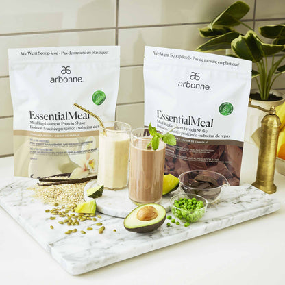 Arbonne EssentialMeal protein shake packs in vanilla and coffee flavors with prepared shakes in glasses, next to a blender and measuring spoons with protein powder.