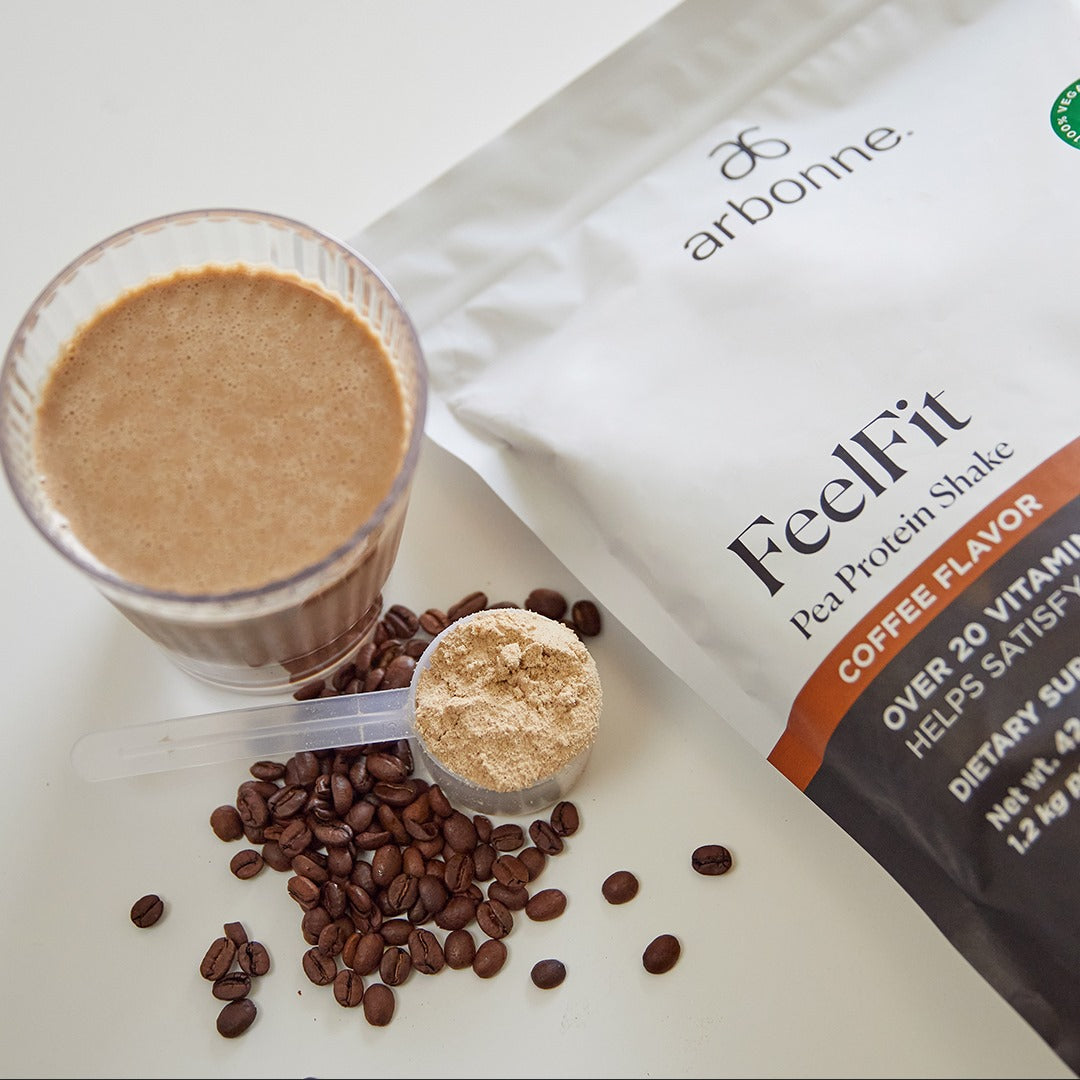 Arbonne FeelFit coffee-flavored pea protein shake in a glass with scattered coffee beans and a measuring scoop.