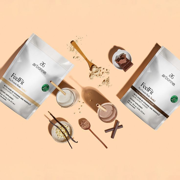 Arbonne FeelFit pea protein shake packs in vanilla and chocolate flavors with prepared shakes, protein powder scoops, vanilla beans, white chocolate chips, and a chocolate bar on a beige background.