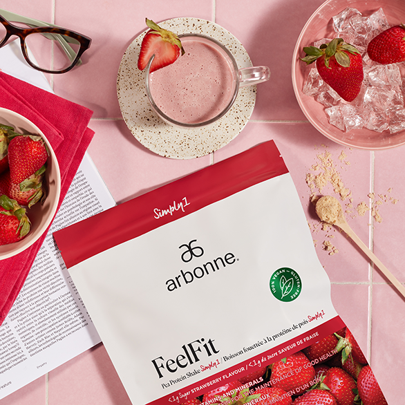 Flat lay of Arbonne FeelFit strawberry protein shake with fresh strawberries, a glass of shake, and reading glasses on a pink tiled surface.