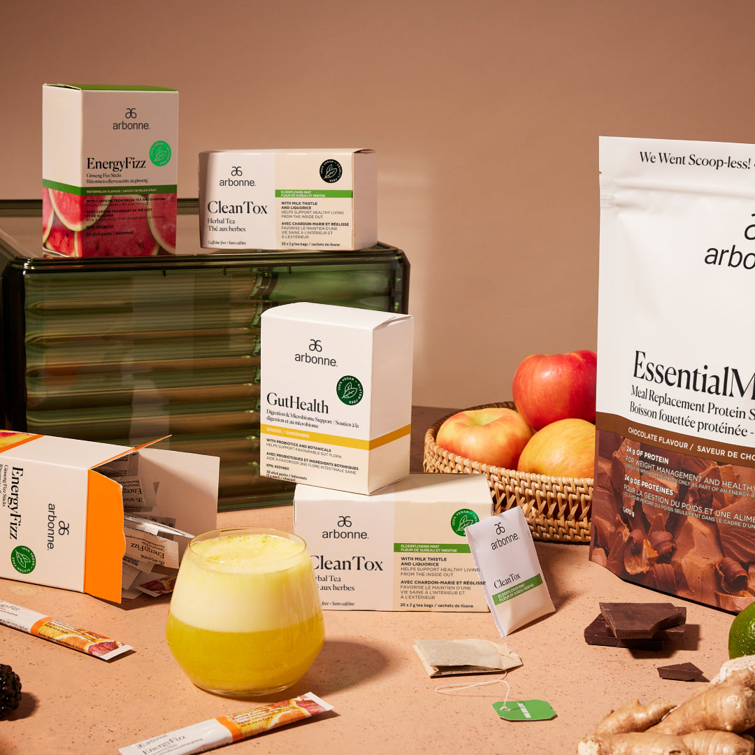 A display of various Arbonne products including EnergyFizz, CleanTox, GutHealth, and EssentialMeal Protein Shake, with a glass of yellow drink and fresh apples in a basket.