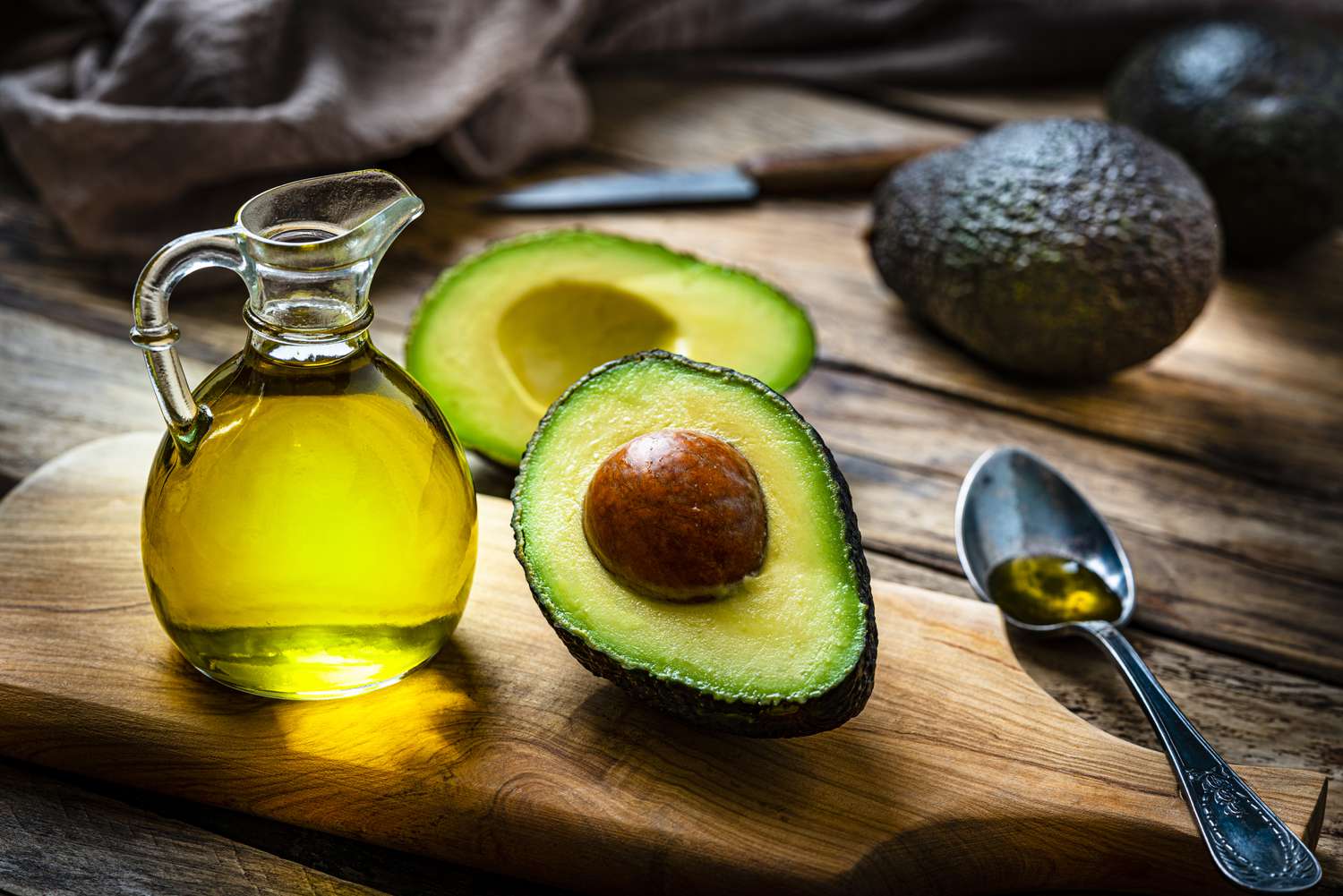 A bottle of avocado oil and a halved avocado on a wooden board, with a spoon of avocado oil beside them.