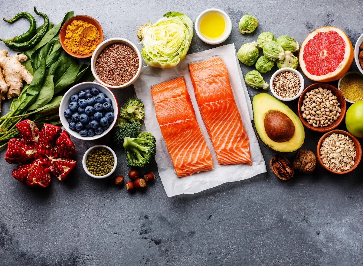 Variety of healthy foods including salmon, avocado, blueberries, pomegranate, grapefruit, Brussels sprouts, broccoli, spinach, nuts, seeds, and grains arranged on a gray surface.