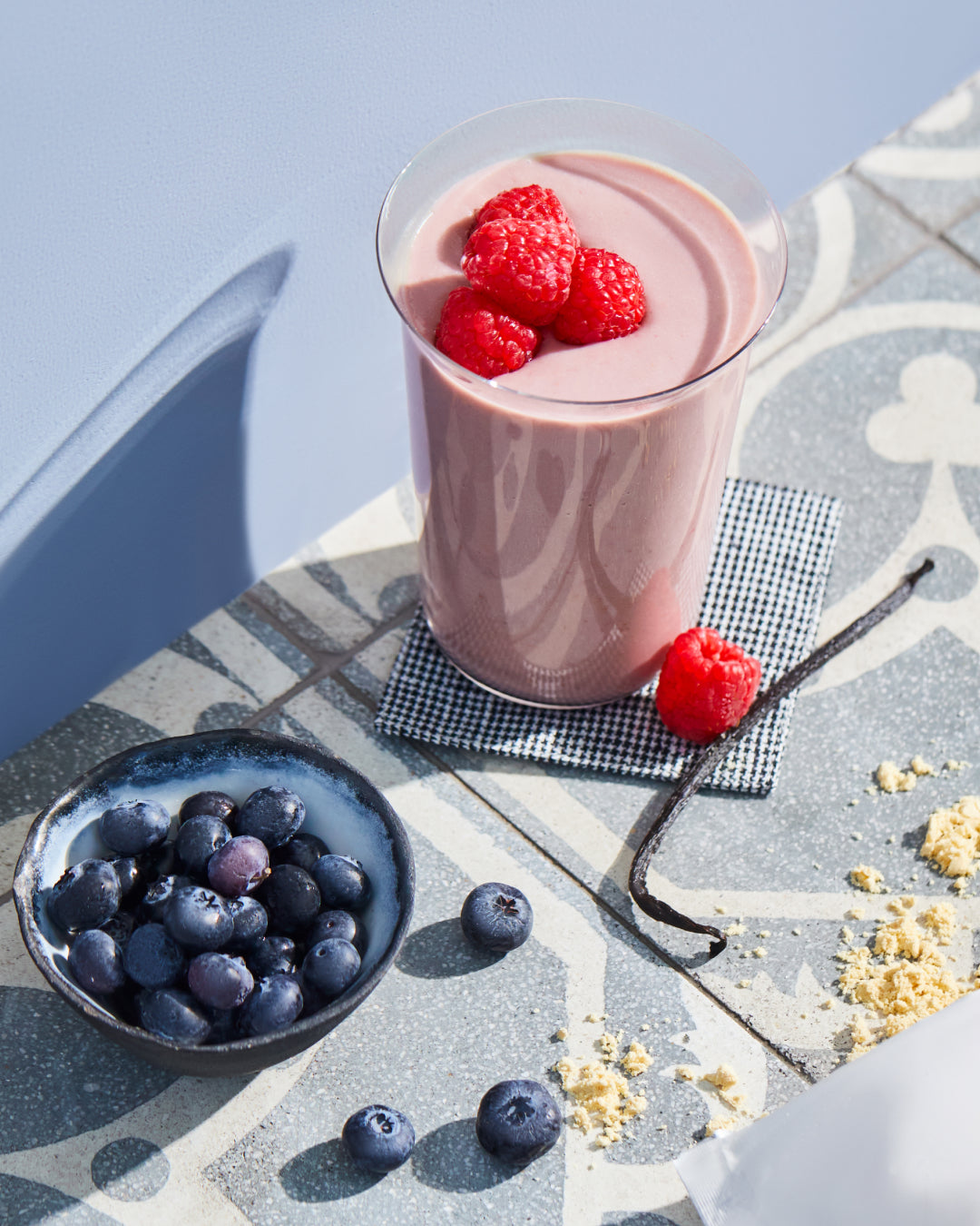 Berry protein shake topped with raspberries beside a bowl of blueberries and vanilla beans on a patterned tile surface