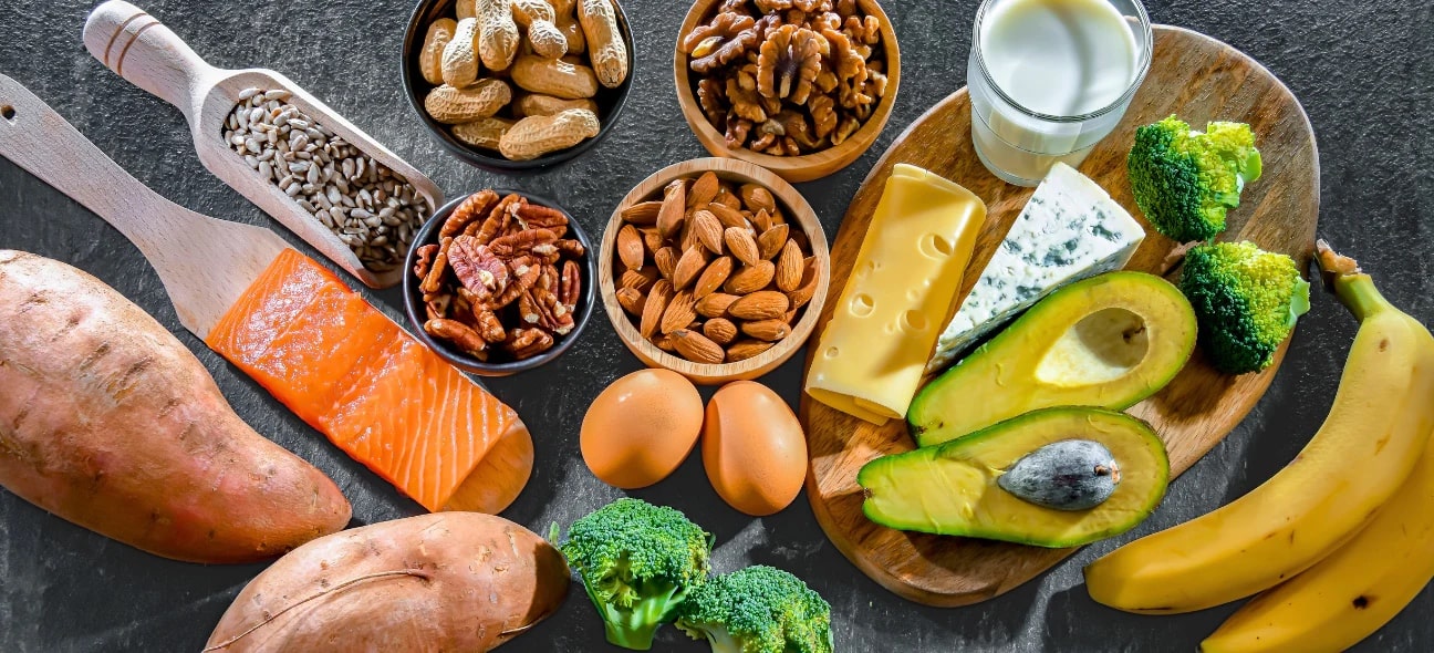 A variety of biotin-rich foods including sweet potatoes, salmon, nuts, seeds, eggs, cheese, avocados, bananas, broccoli, and milk arranged on a dark surface.