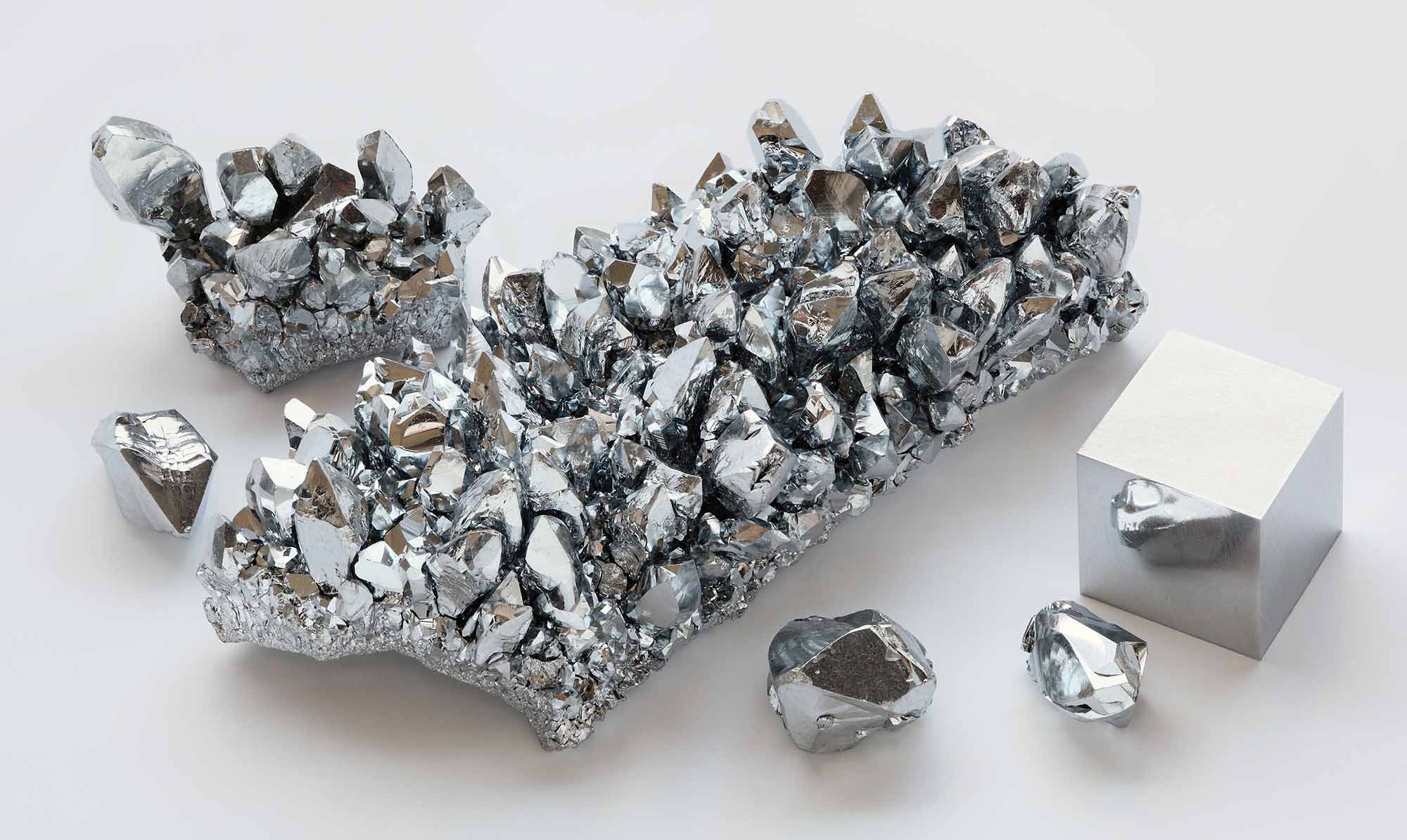 Various forms of chromium, including raw crystals and a polished cube, displayed on a white surface.