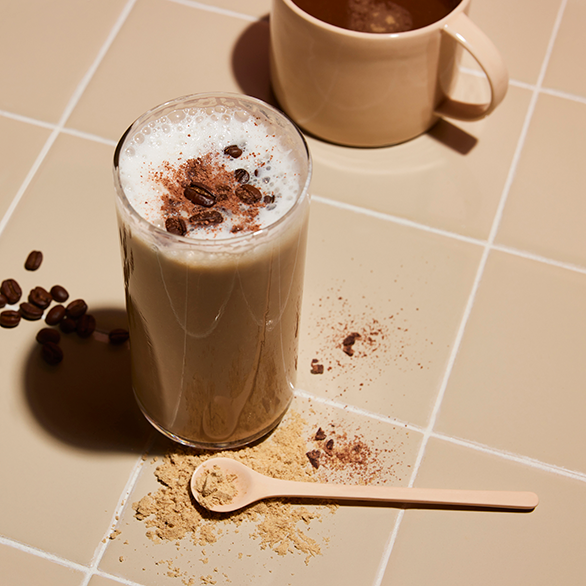 Coffee-flavored protein shake topped with coffee beans, beside a spoonful of protein powder and a cup on a beige tiled counter.