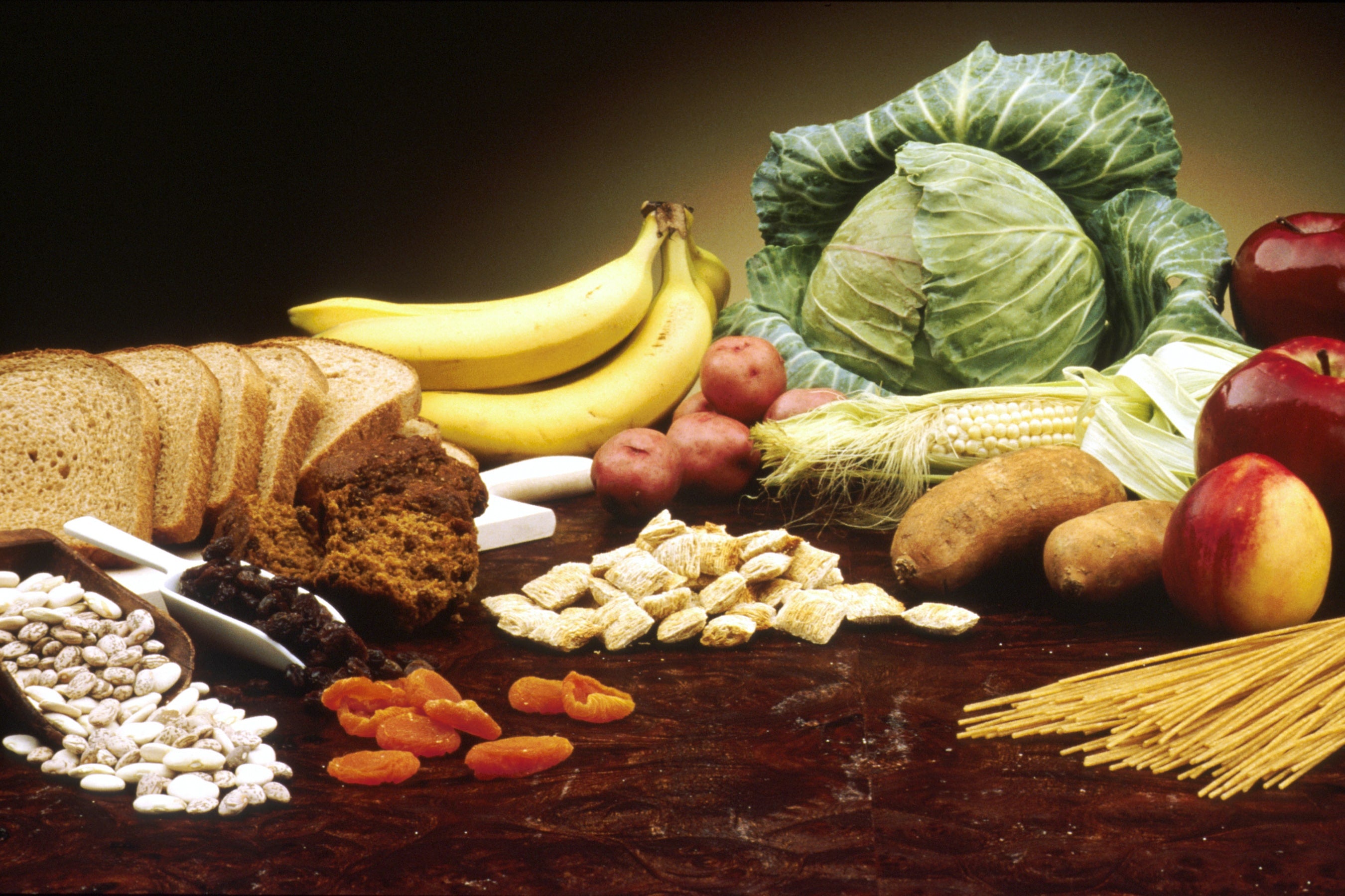 A variety of fiber-rich foods including whole grain bread, muffins, bananas, cabbage, apples, potatoes, corn, dried apricots, beans, raisins, shredded wheat cereal, and spaghetti.