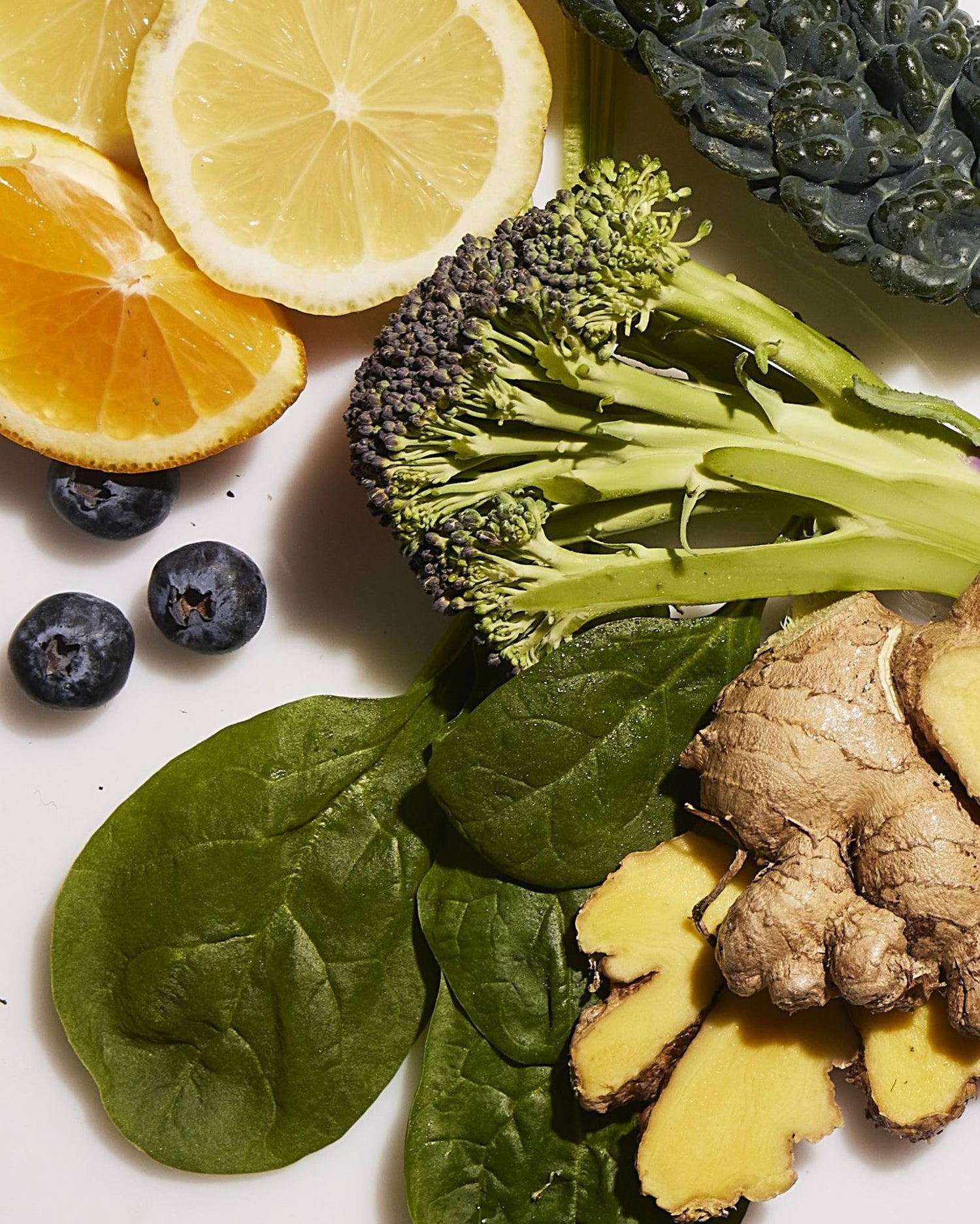 A selection of fresh ingredients including lemon slices, orange slices, broccoli, kale, blueberries, spinach leaves, and ginger root.