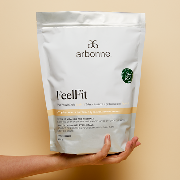 Hand holding a bag of Arbonne FeelFit vanilla pea protein shake against a cream background.