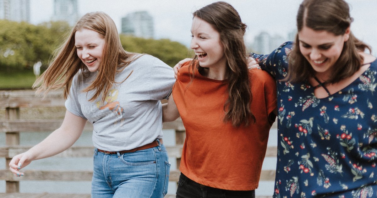 Three young women smiling and laughing together outdoors. They have their arms around each other and appear to be enjoying a fun moment. The background includes trees and a cityscape.