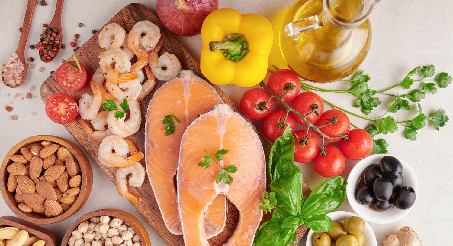 A variety of healthy foods including salmon, shrimp, cherry tomatoes, yellow bell pepper, basil, olives, almonds, chickpeas, and olive oil arranged on a light surface.