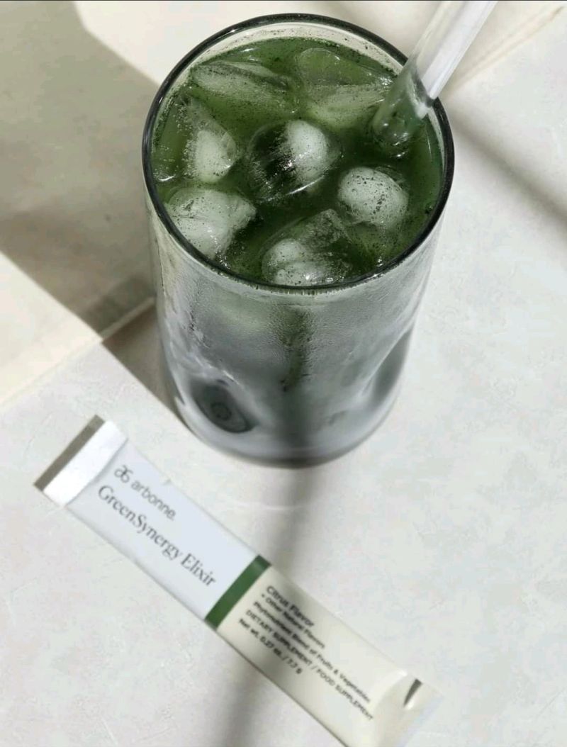 Glass of iced Arbonne GreenSynergy Elixir drink with a straw next to a packet of the elixir.