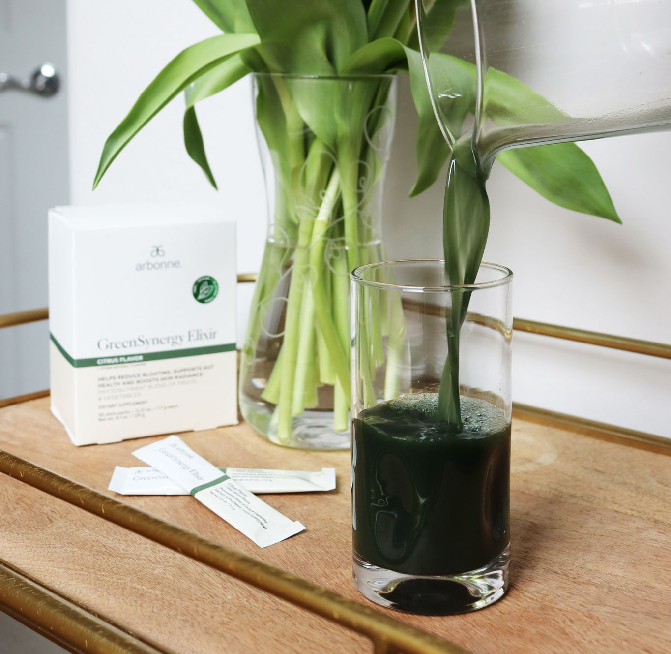 Pouring Arbonne GreenSynergy Elixir into a glass on a wooden table, with the product box and packets nearby, and a vase of green leaves in the background.