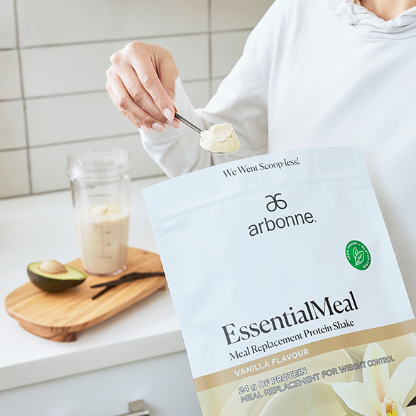 Person preparing a vanilla Arbonne EssentialMeal protein shake with a spoon and packet on kitchen counter.