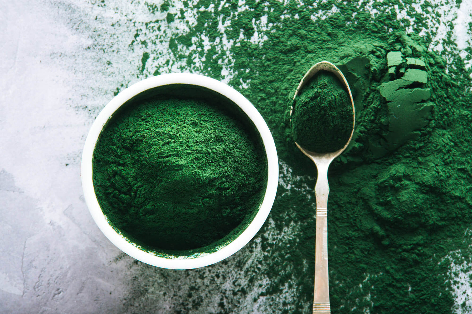  Bowl of vibrant green spirulina powder next to a spoonful of spirulina powder on a textured surface.