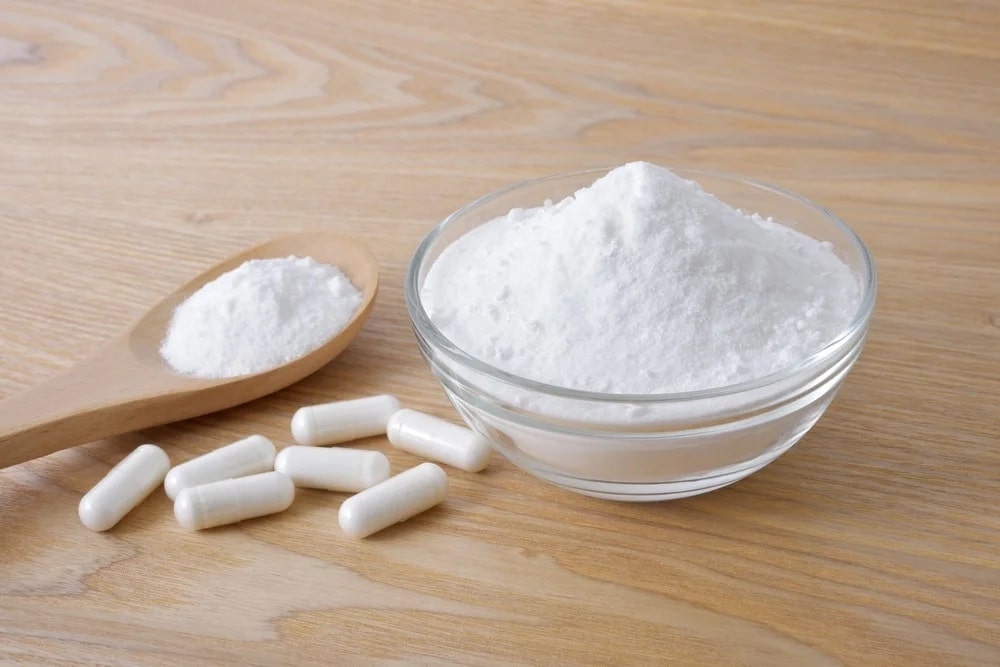 A bowl of white Vegan Taurine powder, a wooden spoon with powder, and white capsules on a wooden surface.