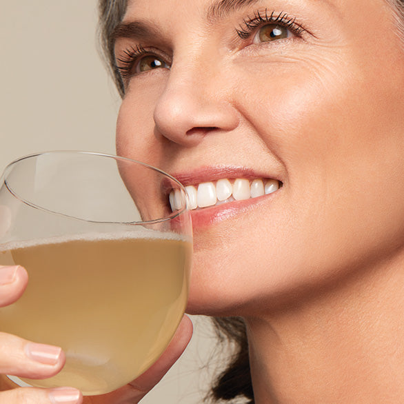 Smiling woman enjoying a sip of Arbonne natural gut health drink, holding a clear glass with golden liquid.