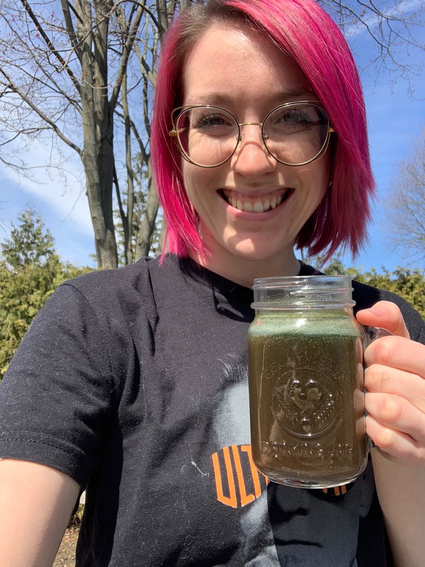 A smiling woman with pink hair and glasses is holding a mason jar filled with a green drink called Green Gut Glow (GGD) by Arbonne. The woman is wearing a black t-shirt and standing outdoors with trees in the background.